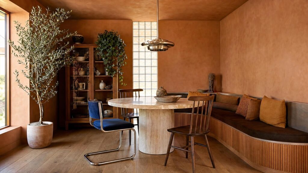 Colour-drenched terracotta dining nook with a travertine table, mismatched dining chairs, glass brick wall and curved bench seat